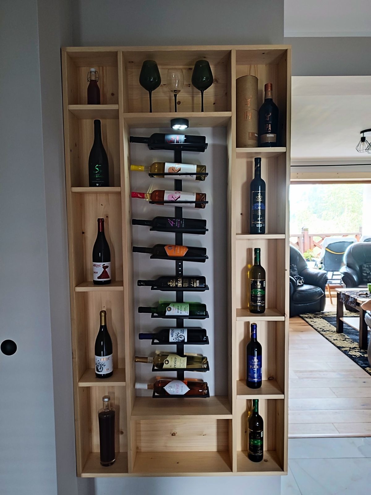 Drewniana szafka na wino z półkami i metalowym uchwytem na butelki, prezentująca różnorodne butelki wina i kieliszki, z widokiem na salon w tle.