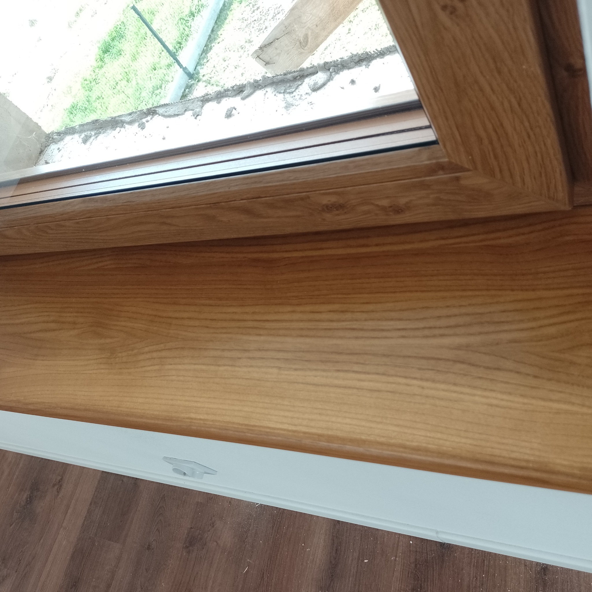 Fragment drewnianego parapetu wewnętrznego z widoczną strukturą drewna, zamontowanego przy oknie z widokiem na zewnątrz, wykończonego białym elementem dolnym.