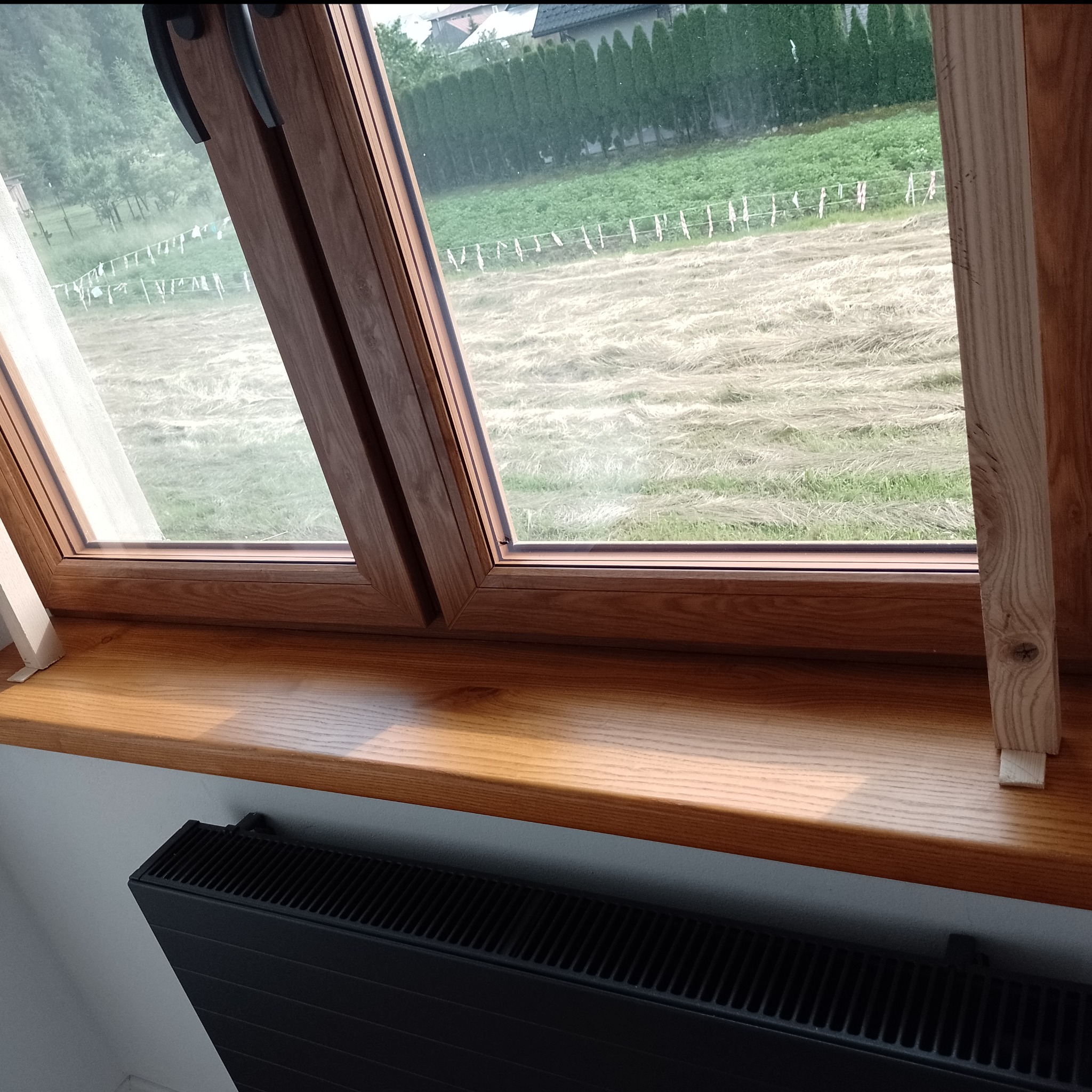 Drewniany parapet pod oknem dachowym z widokiem na łąkę i suszące się pranie, z grzejnikiem pod spodem.