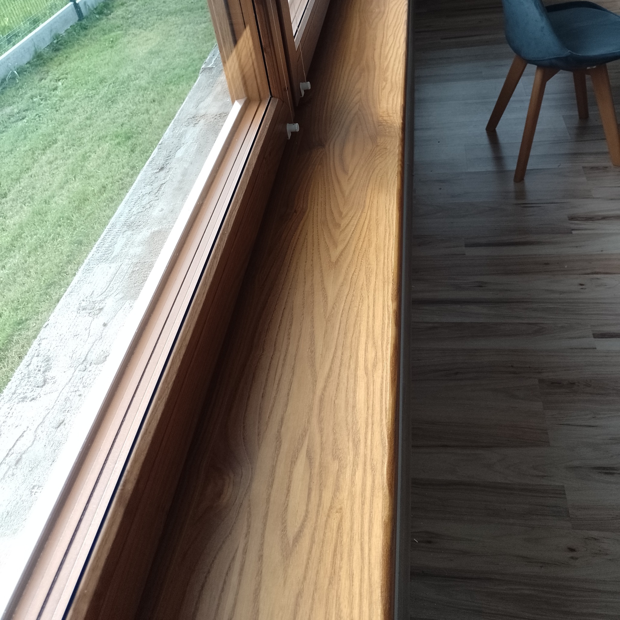 Jasny, drewniany parapet wewnętrzny z widocznym usłojeniem drewna, zamontowany w oknie z widokiem na trawnik, obok fragment podłogi i krzesła.