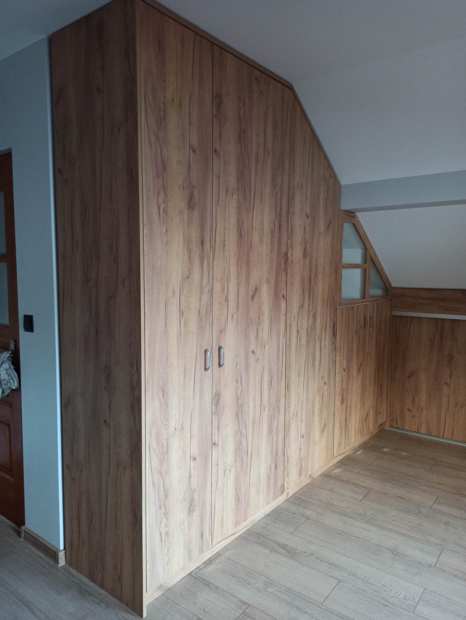 Wysoka zabudowa szafy z drewnopodobną okleiną, dopasowana do skosu poddasza, z widocznymi metalowymi uchwytami i fragmentem podłogi z paneli.