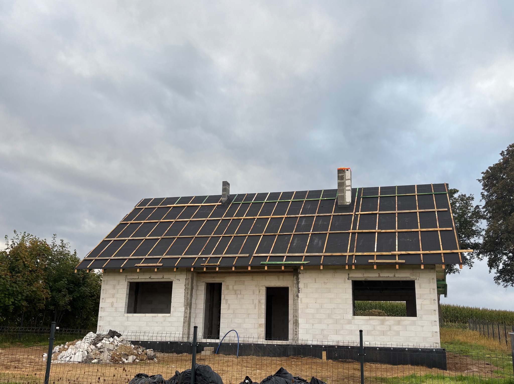 Surowy dom z białego pustaka, czarną membraną i drewnianą łatą na dachu, ogrodzony siatką. Widoczne gruz i worki. Pochmurne niebo w tle.