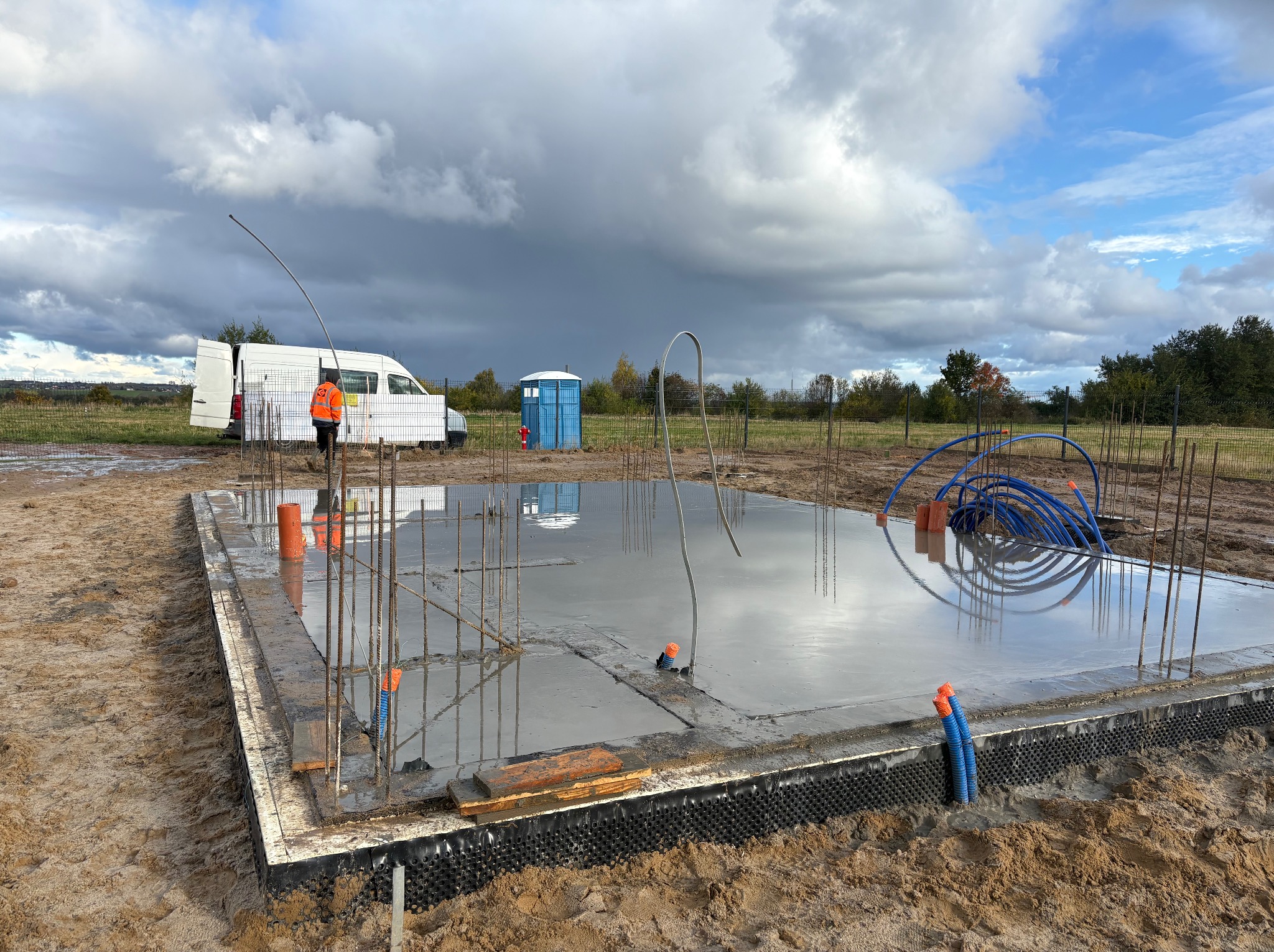 Wylana płyta fundamentowa z wystającymi prętami zbrojeniowymi i rurami instalacyjnymi. W tle biały bus, toaleta przenośna i pracownik w pomarańczowej kamizelce.