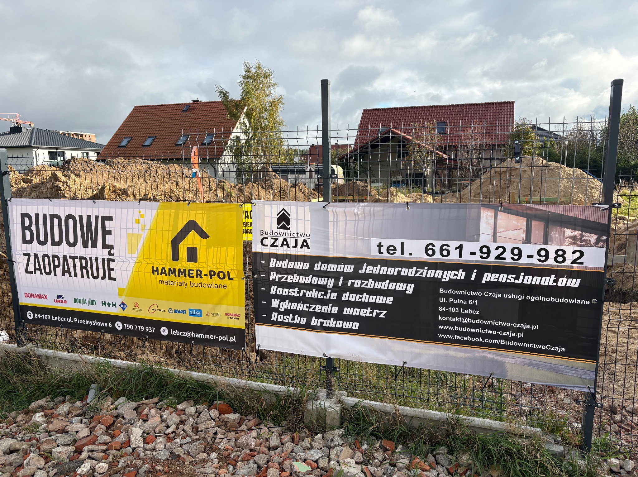 Ogrodzenie placu budowy z banerami firm Hammer-Pol i Budownictwo Czaja, oferujących materiały budowlane i usługi ogólnobudowlane, w tle domy w trakcie budowy.