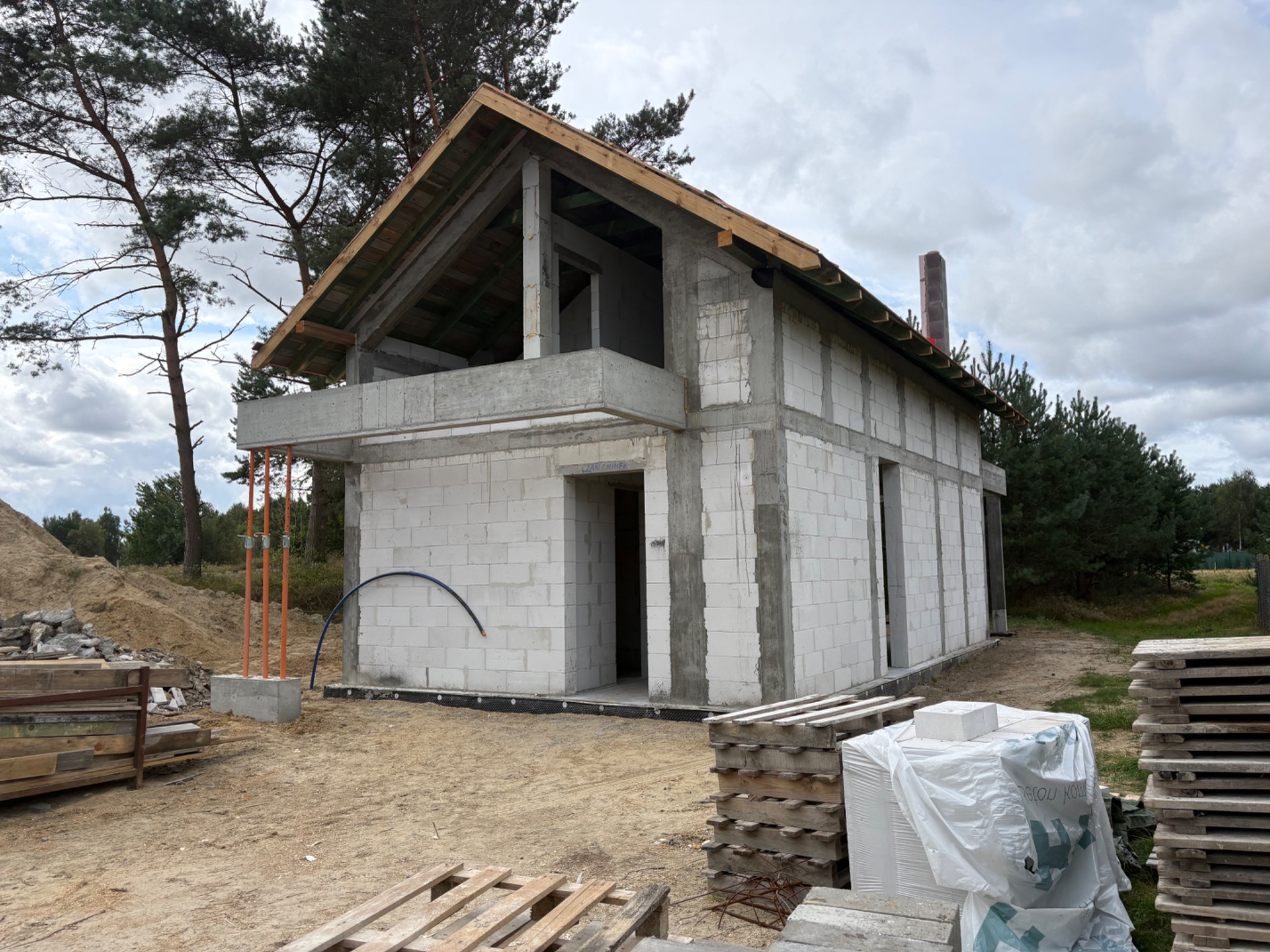 Surowy stan domu z białego bloczka w Łebcz. Widoczne niezadaszone ściany, komin i elementy konstrukcji dachu. Na pierwszym planie palety i materiały budowlane.