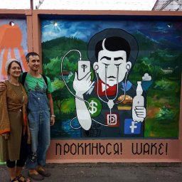 Mykhailo Lapshyn - Para pozuje przed muralem przedstawiającym postać z podłączonym telefonem, płaczącą ikonami aplikacji i trzymającą butelkę, na tle górskiego krajobrazu.