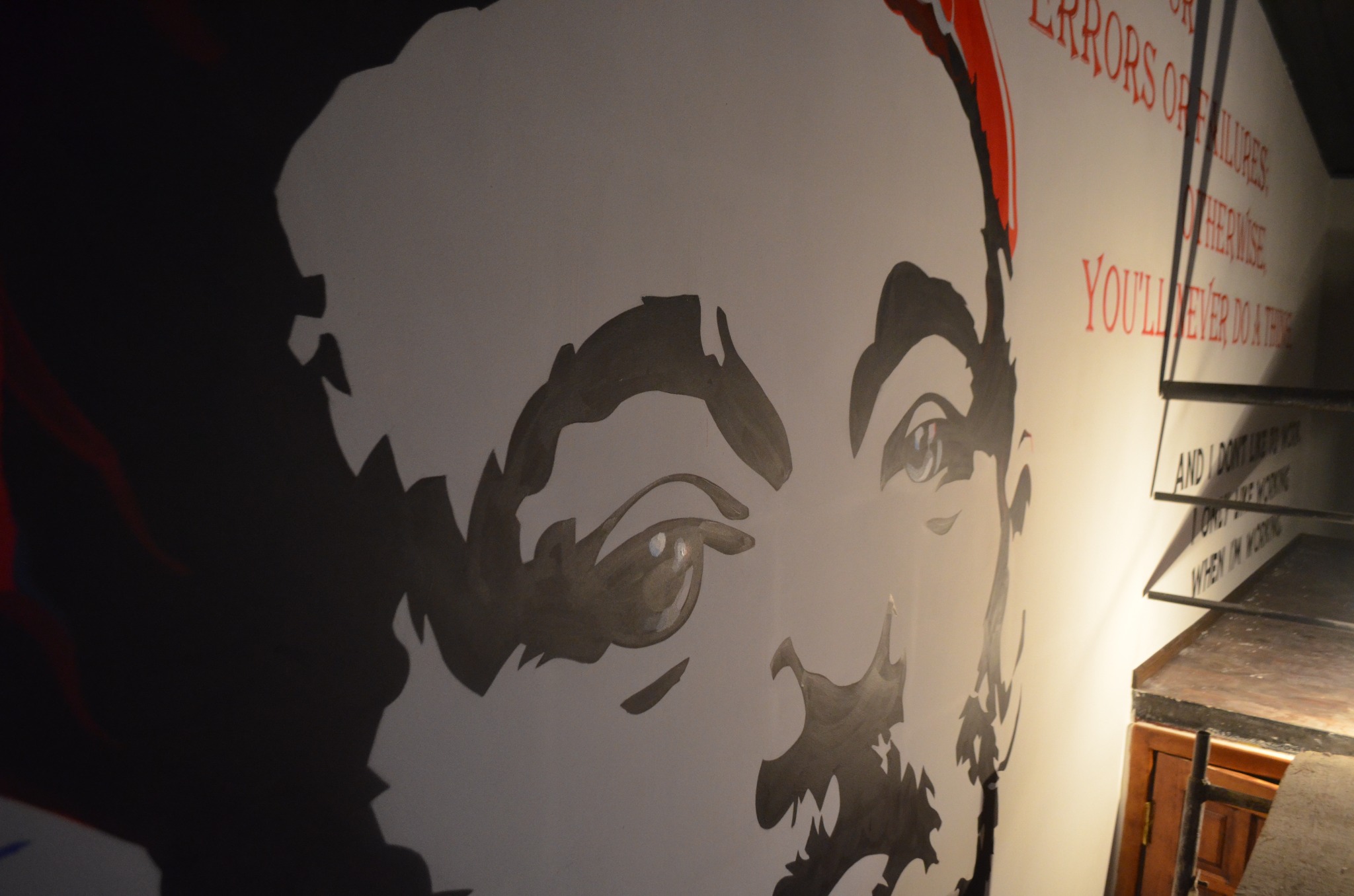 Czarno-biały portret twarzy z czerwonymi akcentami i napisem na ścianie, widoczny z boku w słabym oświetleniu.