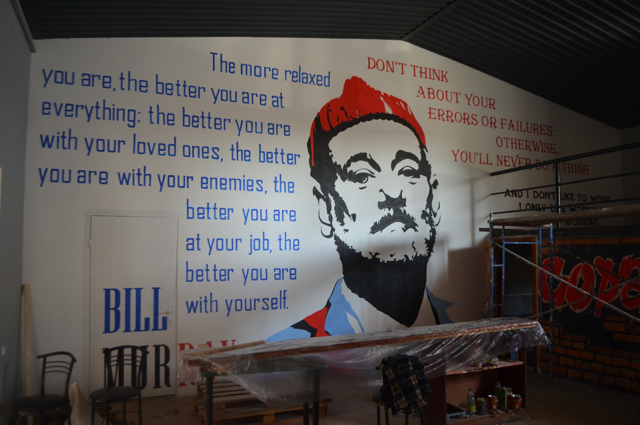 Ściana z muralem przedstawiającym portret w stylu pop-art i motywacyjne cytaty, wykonanym w odcieniach niebieskiego, czerwonego i czarnego.