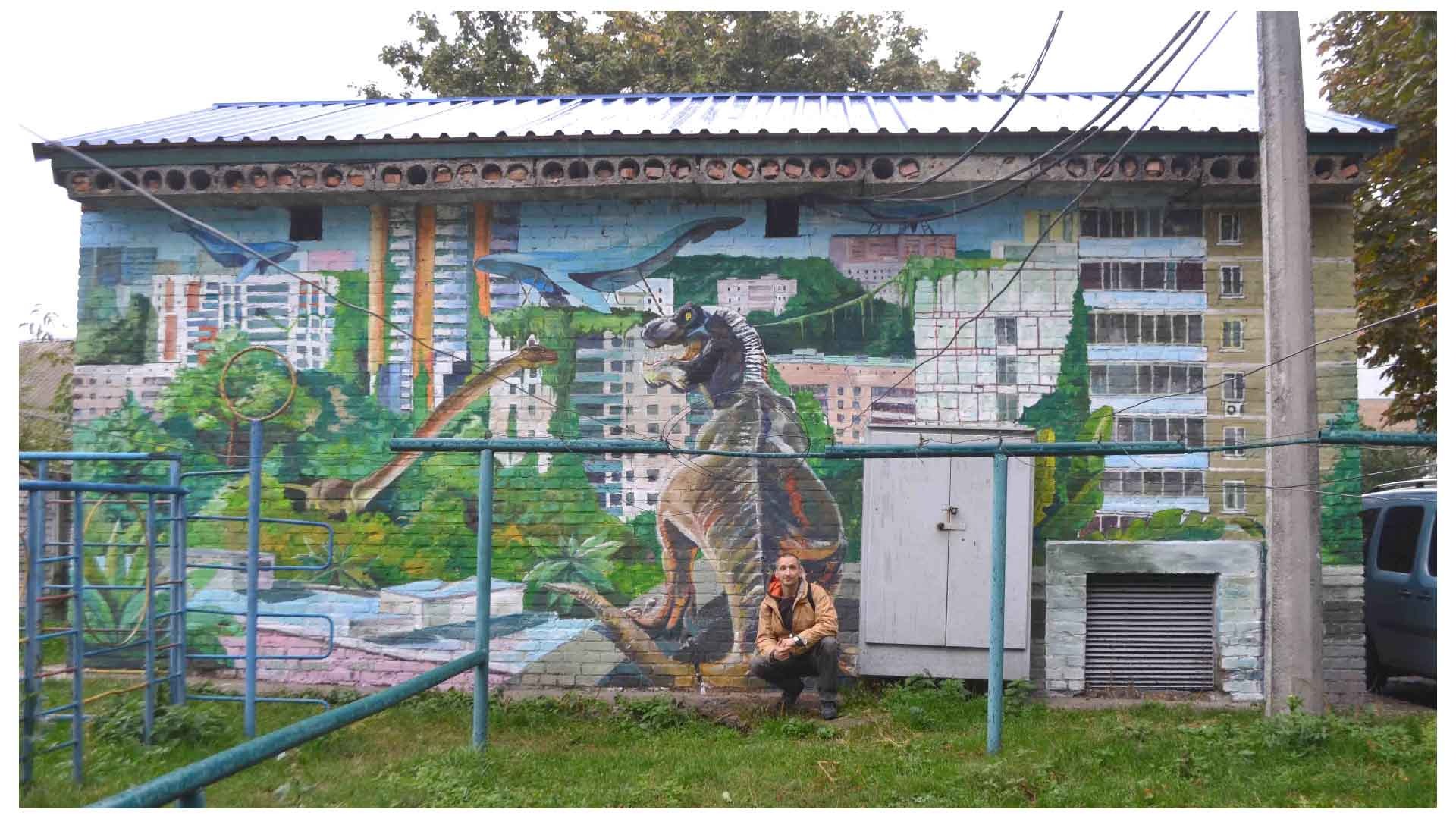Mural na ścianie budynku przedstawiający dinozaury w miejskim krajobrazie, z mężczyzną pozującym na tle obrazu.