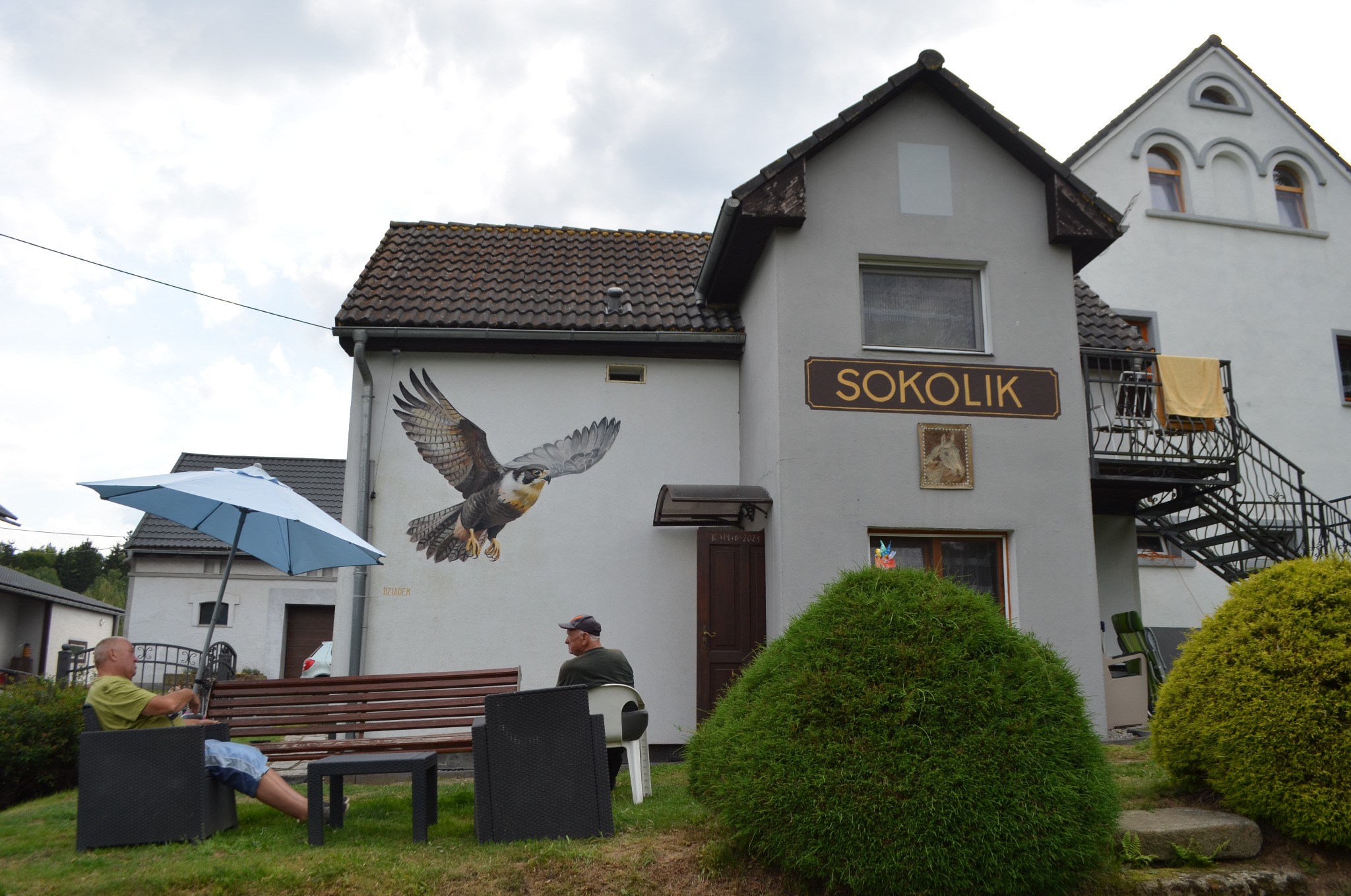 Budynek z muralem przedstawiającym sokoła w locie, obok siedzący mężczyzna na krześle, widoczny napis 'SOKOLIK' na budynku.