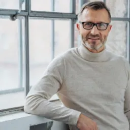 Portret mężczyzny w okularach, opierającego się o parapet okna z podzielonymi szybami, w jasnym swetrze z golfem.