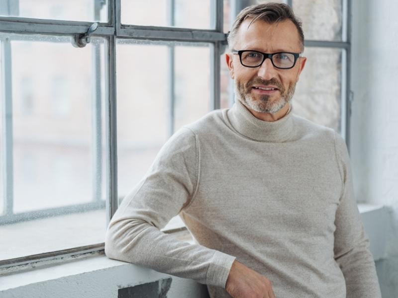 Portret mężczyzny w okularach, opierającego się o parapet okna z podzielonymi szybami, w jasnym swetrze z golfem.