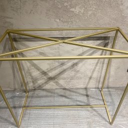 METAL-ART MICHAŁ BUCZYŃSKI - Złota, metalowa konstrukcja ramy stołu z krzyżowym wzmocnieniem, ustawiona na folii bąbelkowej, na tle szarej, teksturowanej ściany.