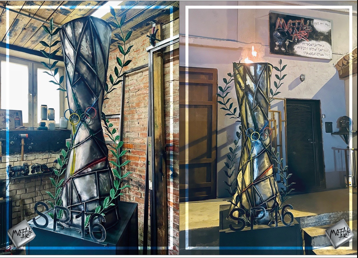 Metalowa rzeźba w Szczecinie, przypominająca znicz olimpijski z płomieniem, otoczona metalową girlandą z liści, z widocznymi detalami warsztatu w tle.