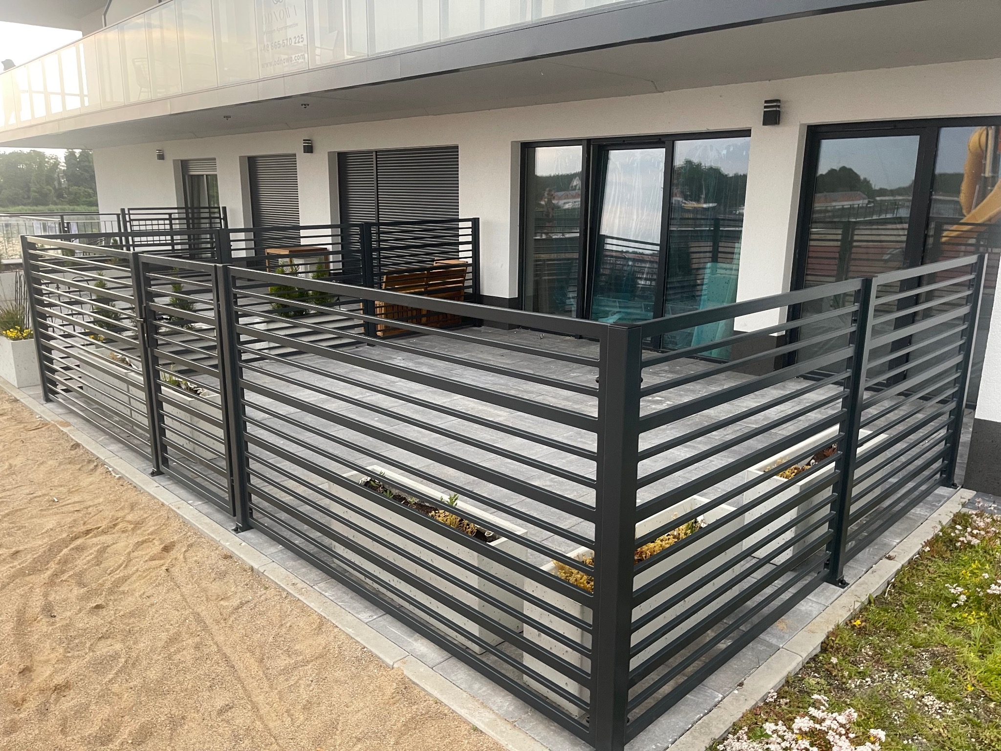 Nowoczesna, pozioma, metalowa konstrukcja oddzielająca balkon od otoczenia, z widocznymi donicami z roślinami, na tle białej elewacji budynku z dużymi oknami.