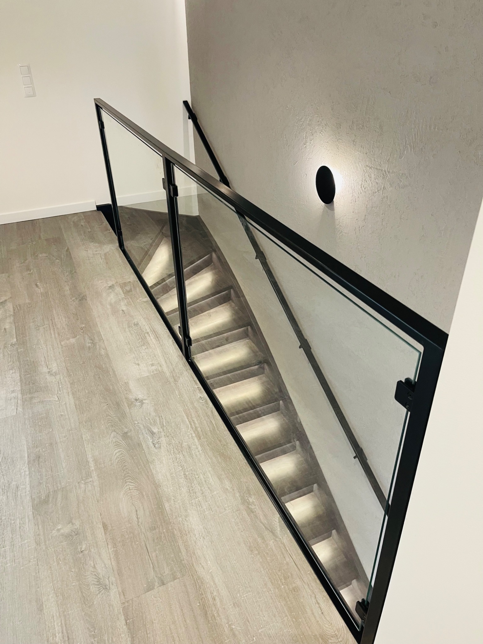 Nowoczesne schody z podświetleniem LED za szklaną balustradą z czarnymi profilami, widok z góry na jasną podłogę i minimalistyczne oświetlenie ścienne.