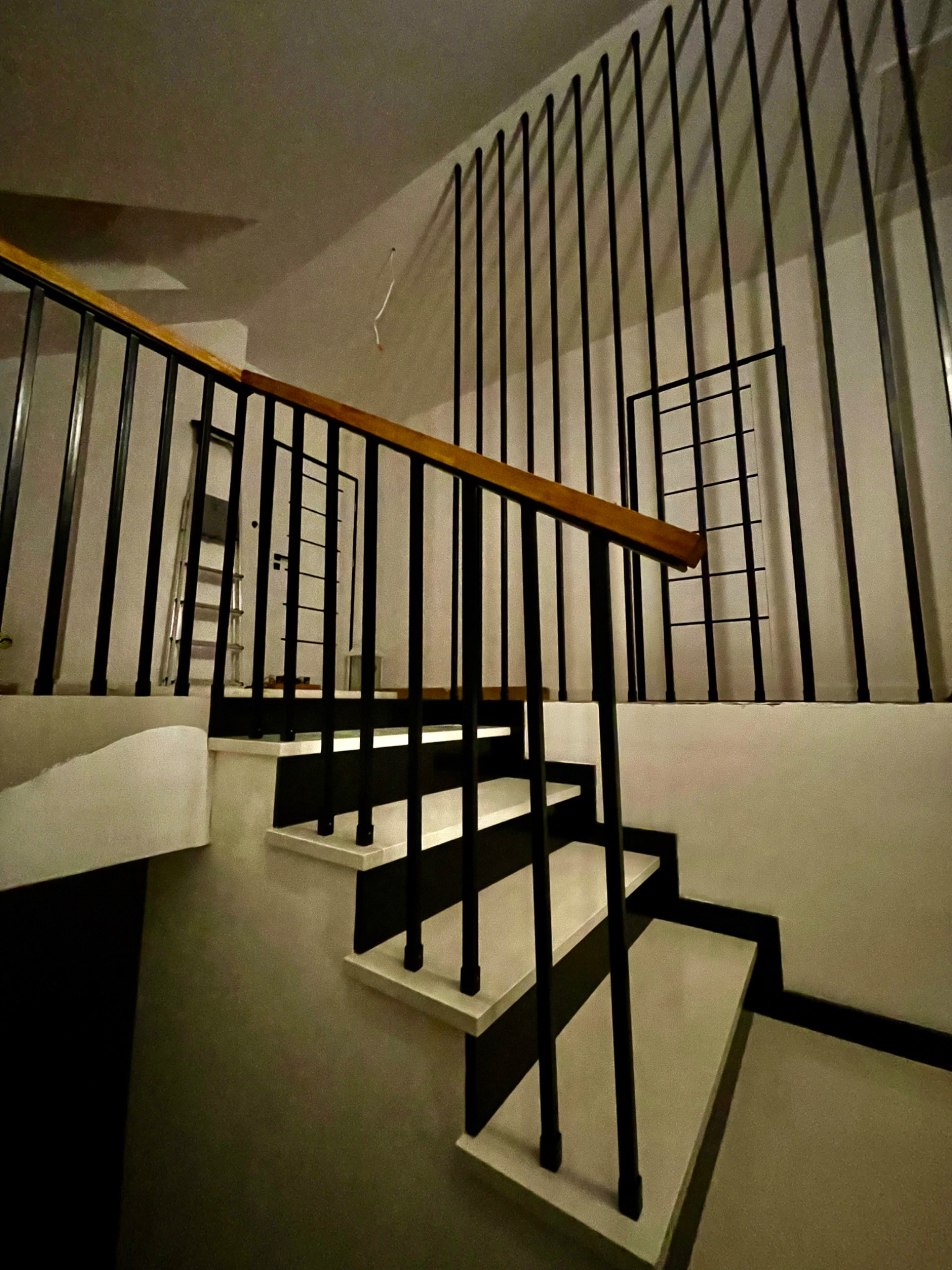 Nowoczesne schody wewnętrzne z czarnymi, pionowymi balustradami, drewnianą poręczą i białymi stopniami z czarnymi krawędziami, widziane z dołu, z drabiną w tle.