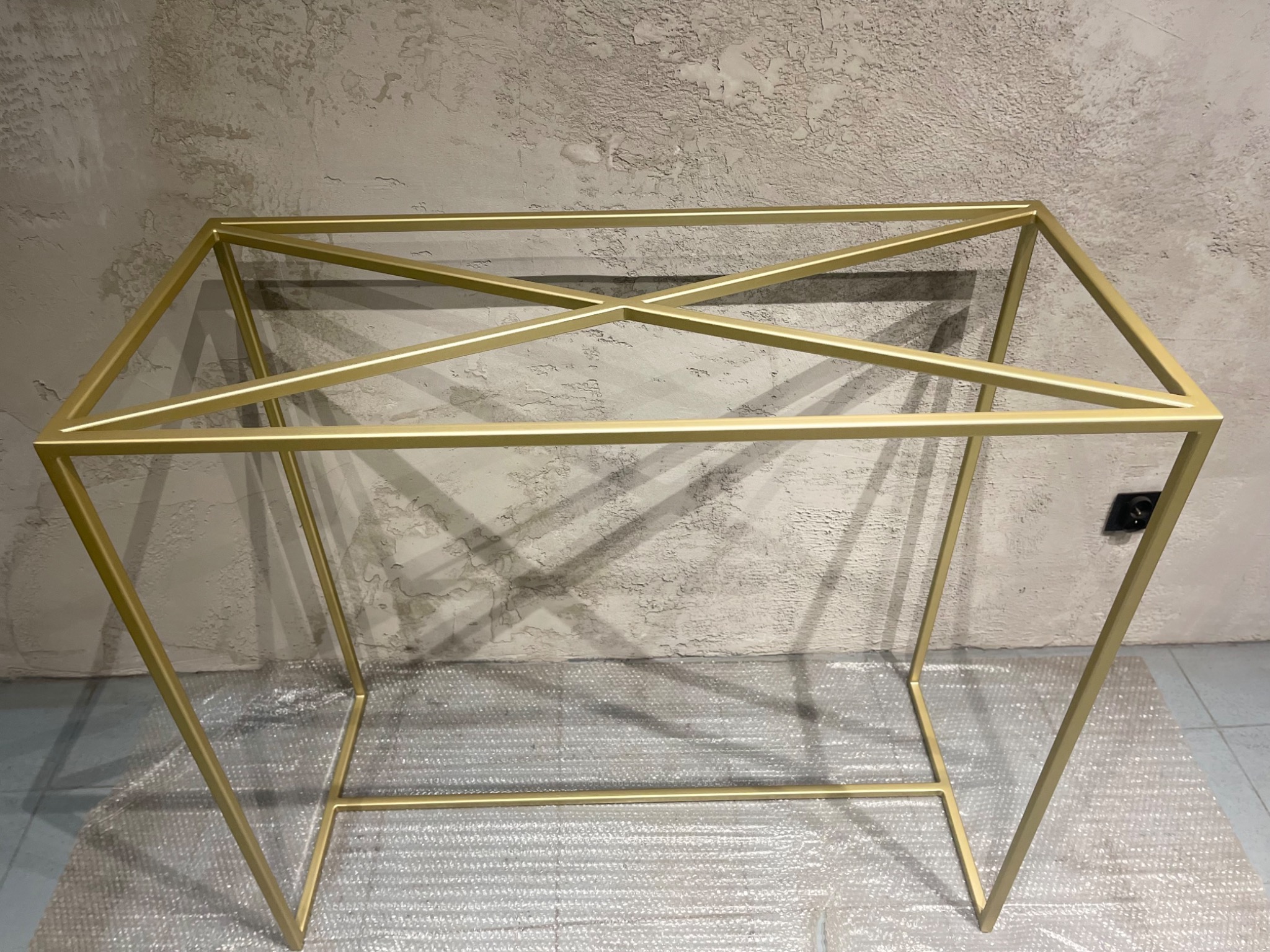 Złota, metalowa konstrukcja ramy stołu z krzyżowym wzmocnieniem, ustawiona na folii bąbelkowej, na tle szarej, teksturowanej ściany.