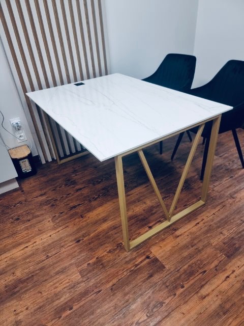 Stół z białym blatem imitującym marmur i złotymi, metalowymi nogami w kształcie litery V, na tle drewnianej ściany i dwóch czarnych krzeseł.