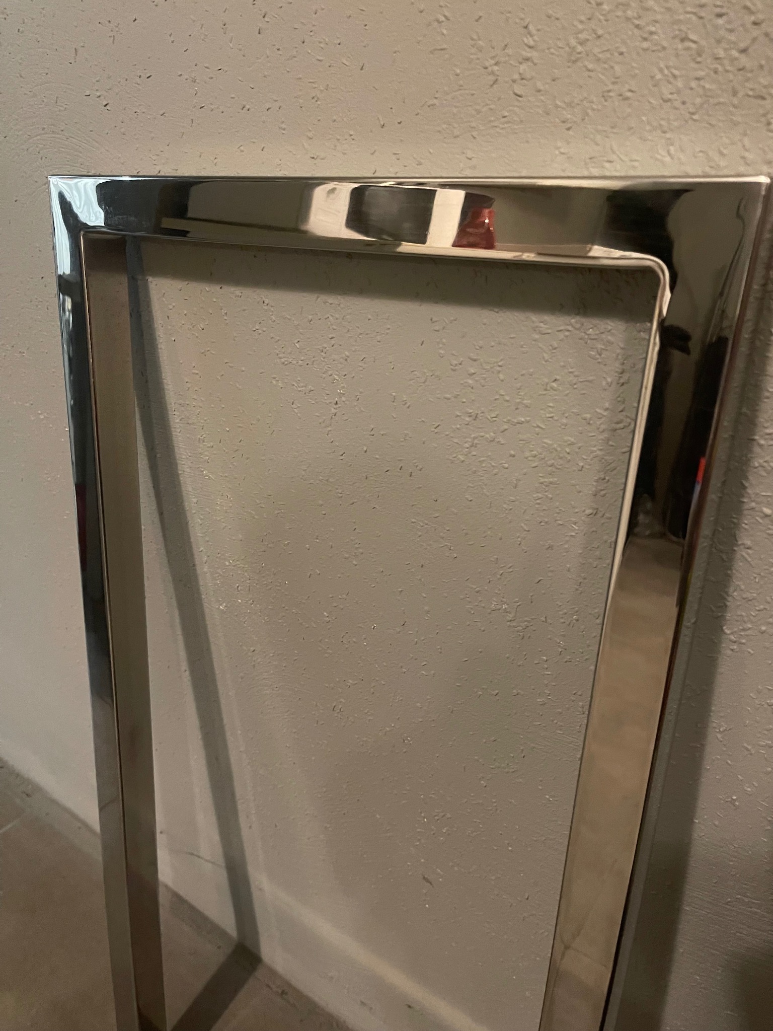 Fragment konstrukcji z polerowanej stali nierdzewnej, prawdopodobnie element ramy lub poręczy, widoczny na tle szarej, teksturowanej ściany.