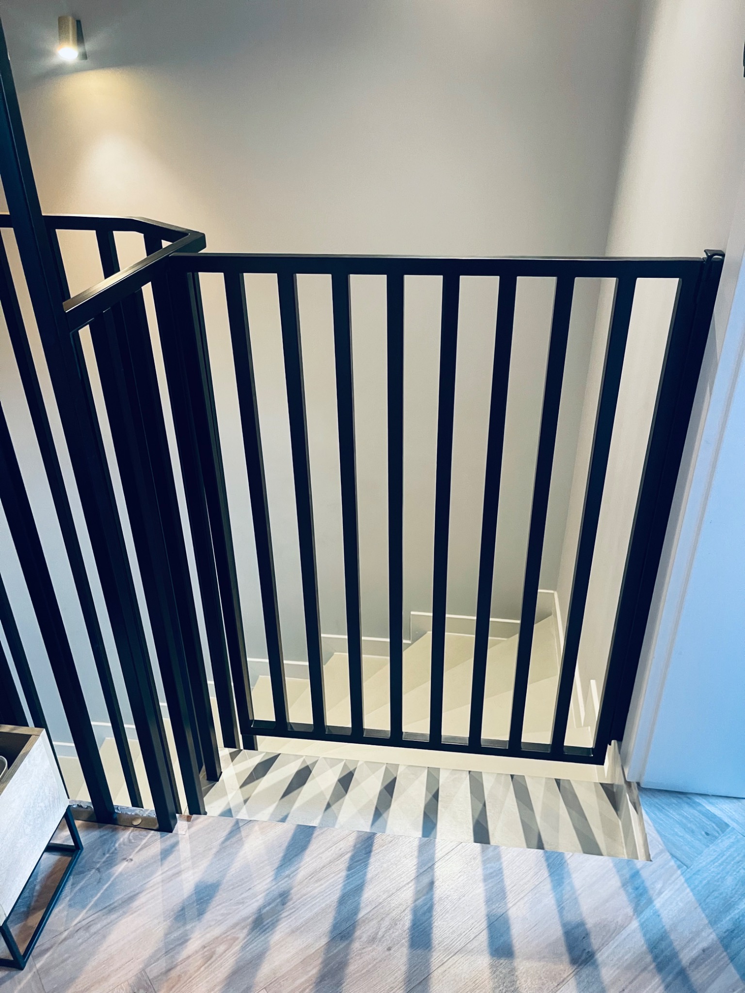 Nowoczesna, czarna barierka schodowa z pionowych elementów, rzucająca cienie na drewnopodobną podłogę i białe schody.