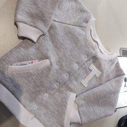 MW - Szara bluza dziecięca z białą literą T i kieszenią z kolorową podszewką, zapinana na zatrzaski, leżąca na jasnym tle.