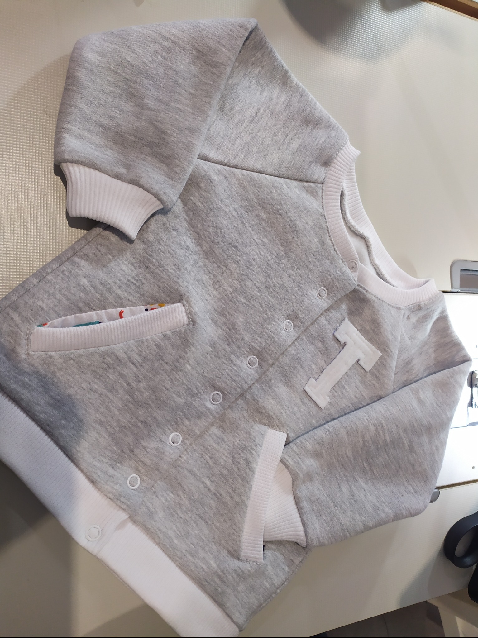 Szara bluza dziecięca z białą literą T i kieszenią z kolorową podszewką, zapinana na zatrzaski, leżąca na jasnym tle.