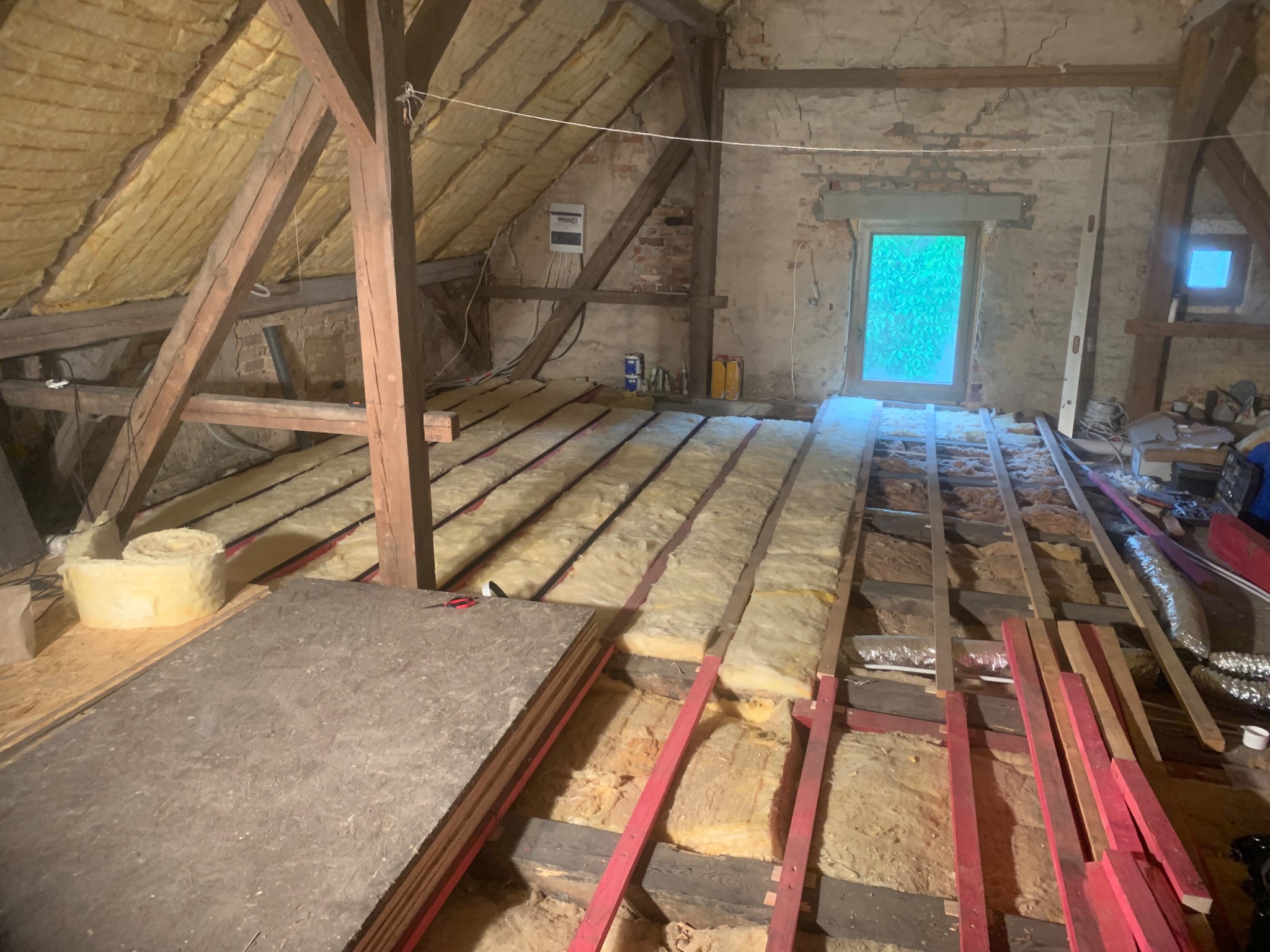 Izolacja poddasza wełną mineralną między drewnianymi legarami, przygotowanie do ułożenia podłogi z płyt drewnopochodnych, widoczne narzędzia i materiały budowlane.