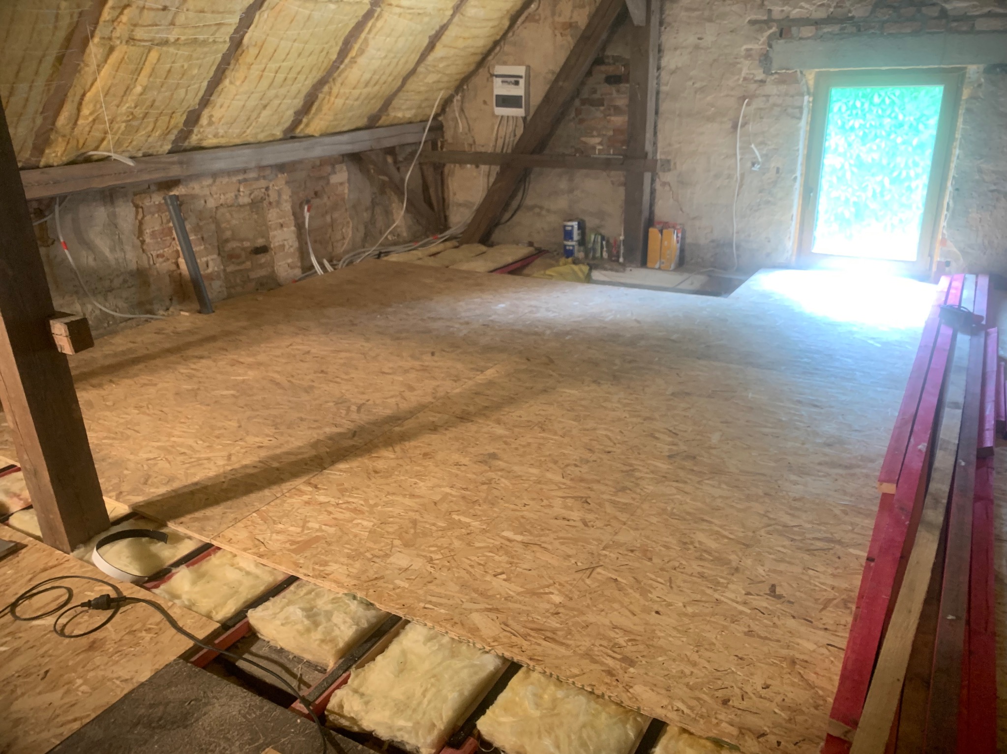 Podłoga na poddaszu w trakcie remontu, płyty OSB ułożone na izolacji z wełny mineralnej, widoczne elementy konstrukcji dachu i ściany z cegły, okno w tle.