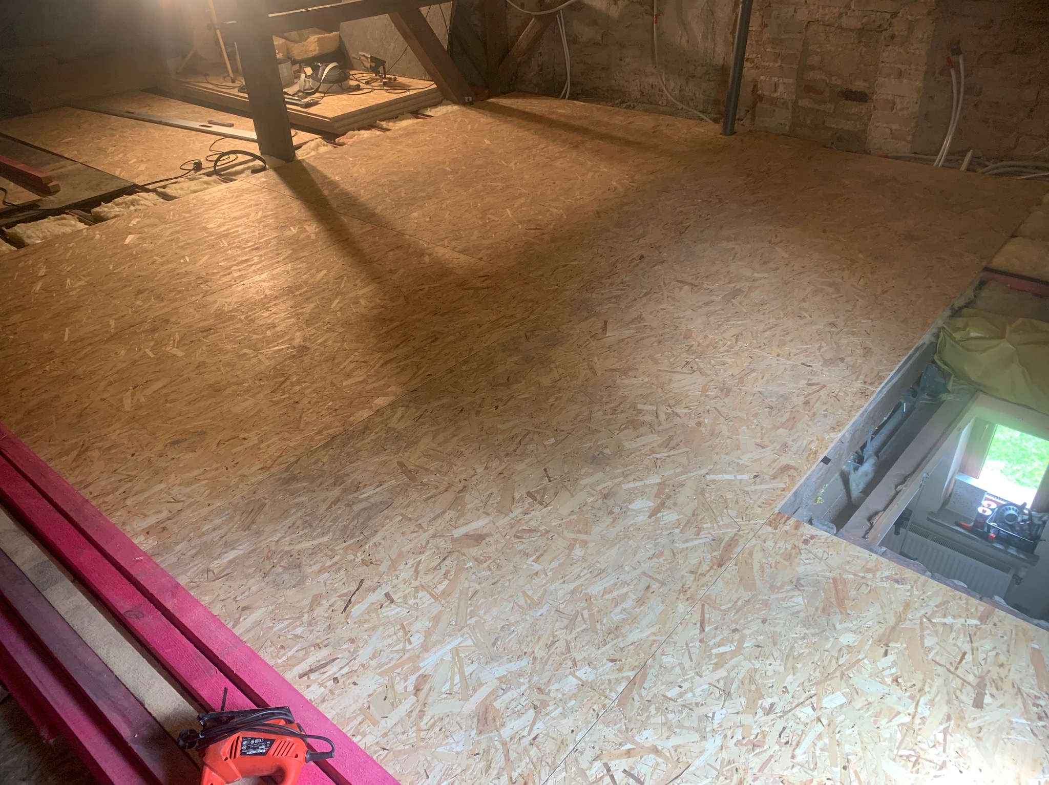 Montaż podłogi z płyt OSB na poddaszu, widoczne ocieplenie między legarami, narzędzia i deski w kolorze fuksji w rogu zdjęcia, surowy stan wykończenia ścian.