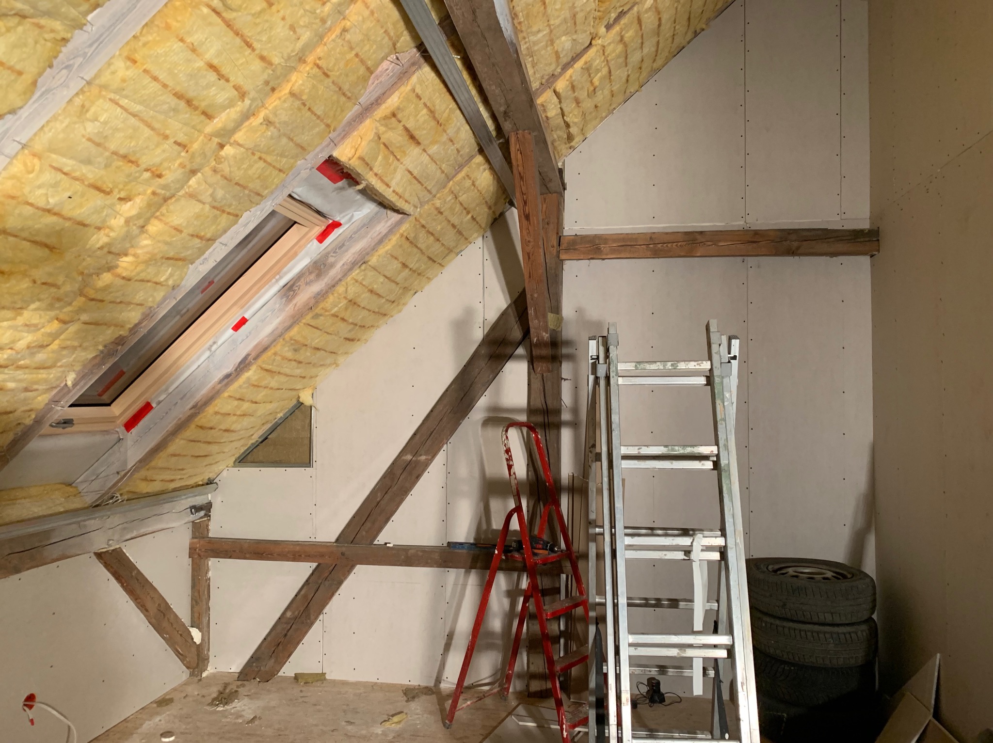 Poddasze w trakcie adaptacji: ocieplenie wełną mineralną widoczne na skośnym dachu, częściowo zamontowane płyty na ścianach, dwie drabiny (czerwona i srebrna) oraz opony samochodowe w rogu...