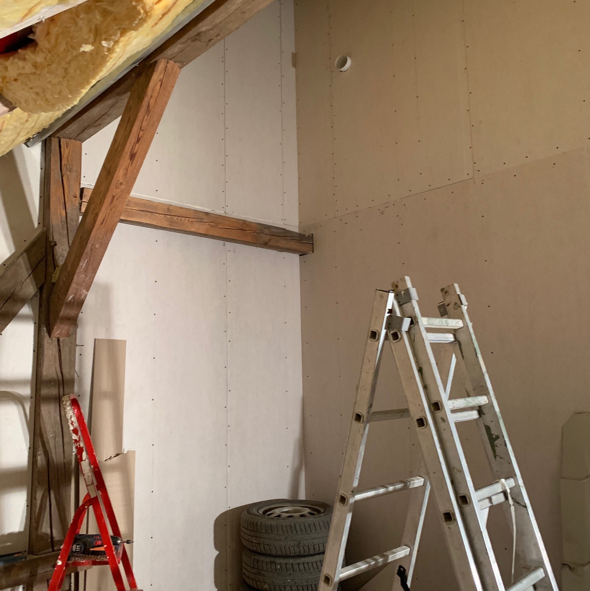 Wnętrze poddasza w trakcie prac wykończeniowych, ściany pokryte płytami z widocznymi wkrętami, drewniane belki stropowe, dwie drabiny (czerwona i aluminiowa) oraz opony samochodowe.