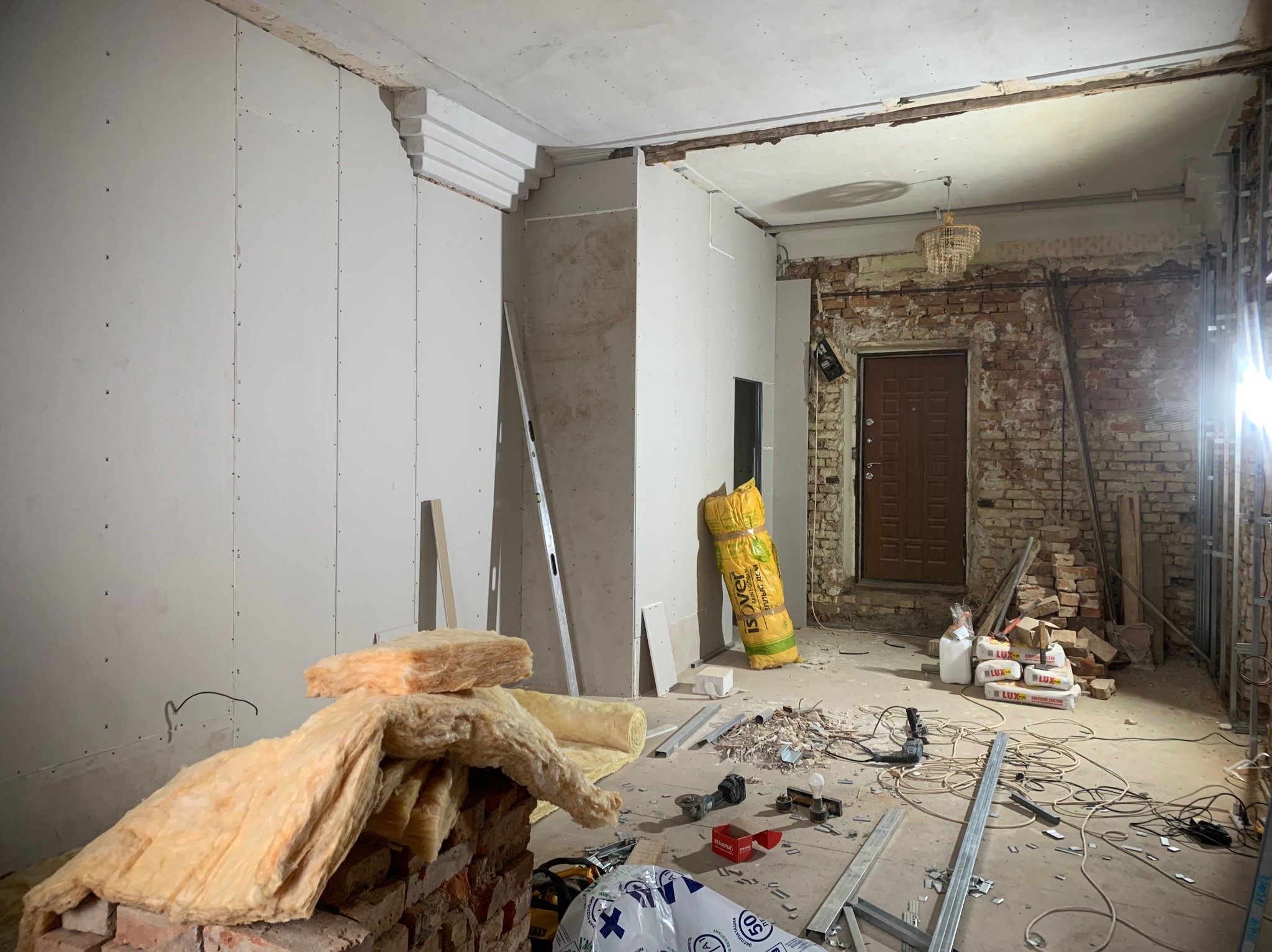 Wnętrze w trakcie remontu: fragment ściany z zamontowanymi płytami, odsłonięta cegła, materiały izolacyjne, narzędzia i worki z zaprawą budowlaną na podłodze.