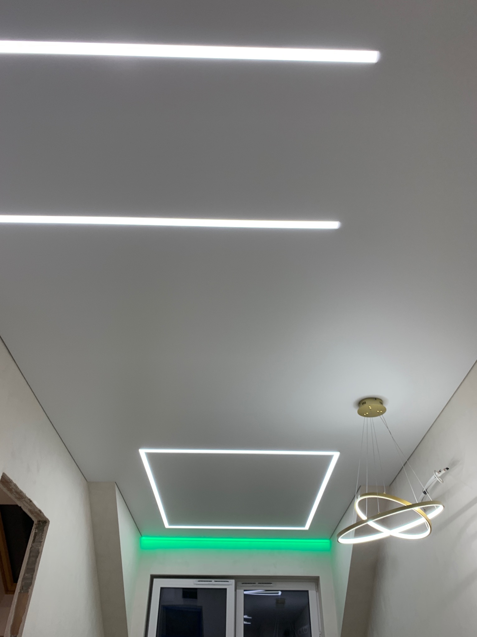 Biały sufit z liniowym oświetleniem LED: dwie długie linie, kwadratowa rama i lampa wisząca z trzema pierścieniami w kolorze złota, nad oknem zielone podświetlenie.