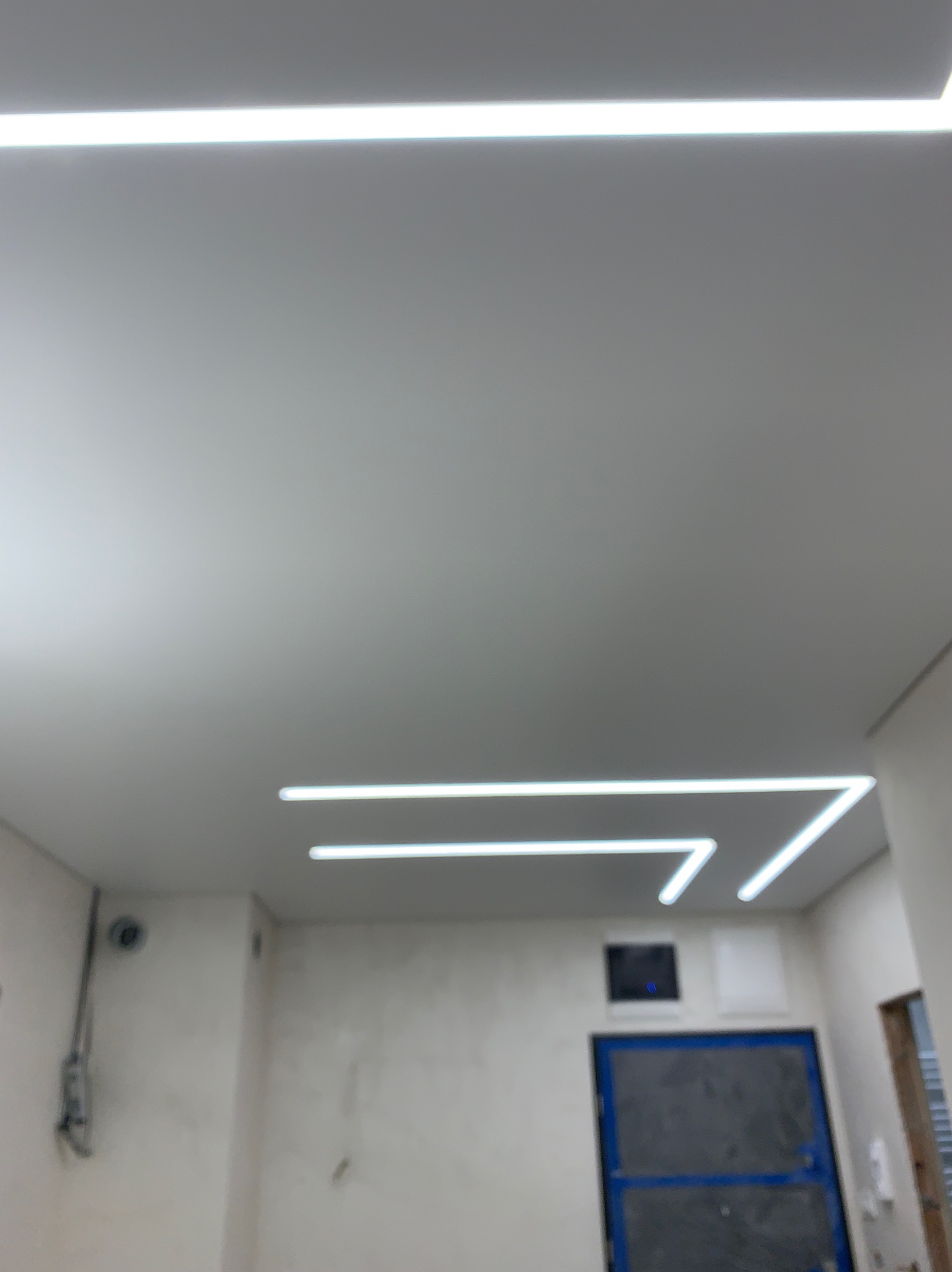 Biały sufit z nowoczesnym oświetleniem LED w kształcie linii i kątów, widoczna szara ściana i niebieskie drzwi w tle.