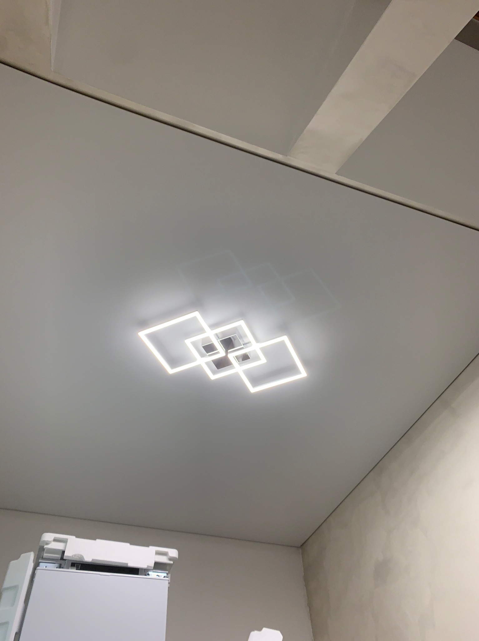 Biały, matowy sufit z nowoczesnym oświetleniem LED w kształcie splecionych kwadratów, widoczne niedoskonałości na ścianie i fragment sprzętu budowlanego w rogu.