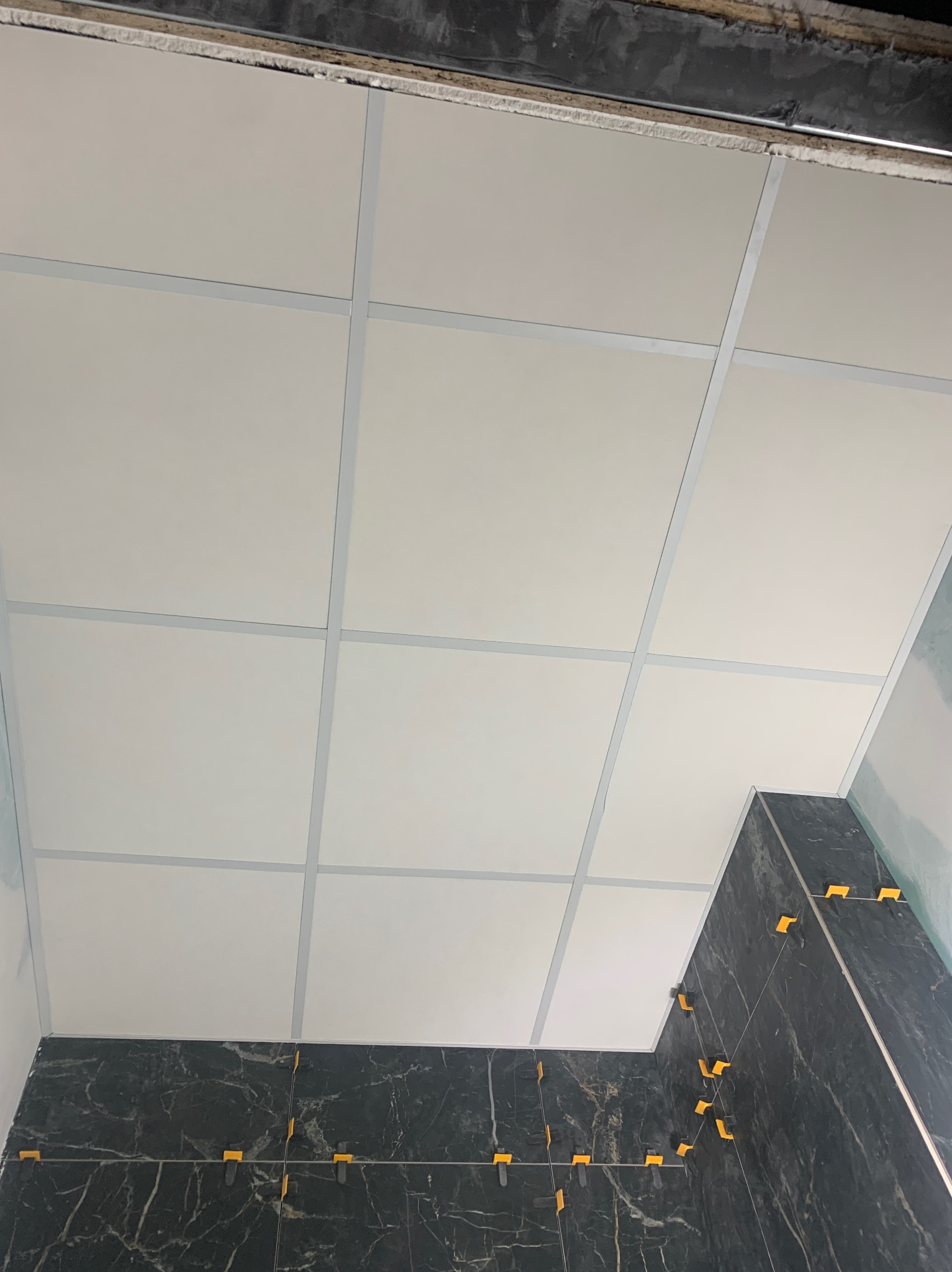 Montaż sufitu podwieszanego z płyt gipsowo-kartonowych w łazience z czarnymi marmurowymi płytkami, widoczne żółte krzyżyki dystansowe.