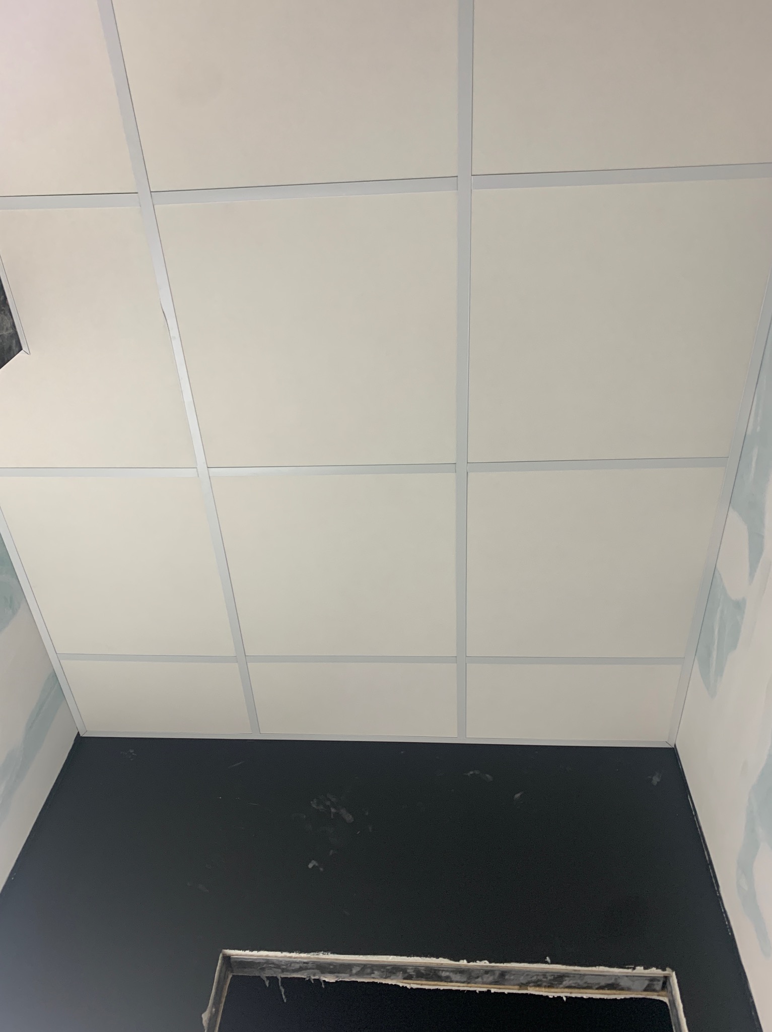 Kwadratowy sufit kasetonowy z widoczną konstrukcją nośną, czarna ściana poniżej i ślady prac wykończeniowych na ścianach bocznych.