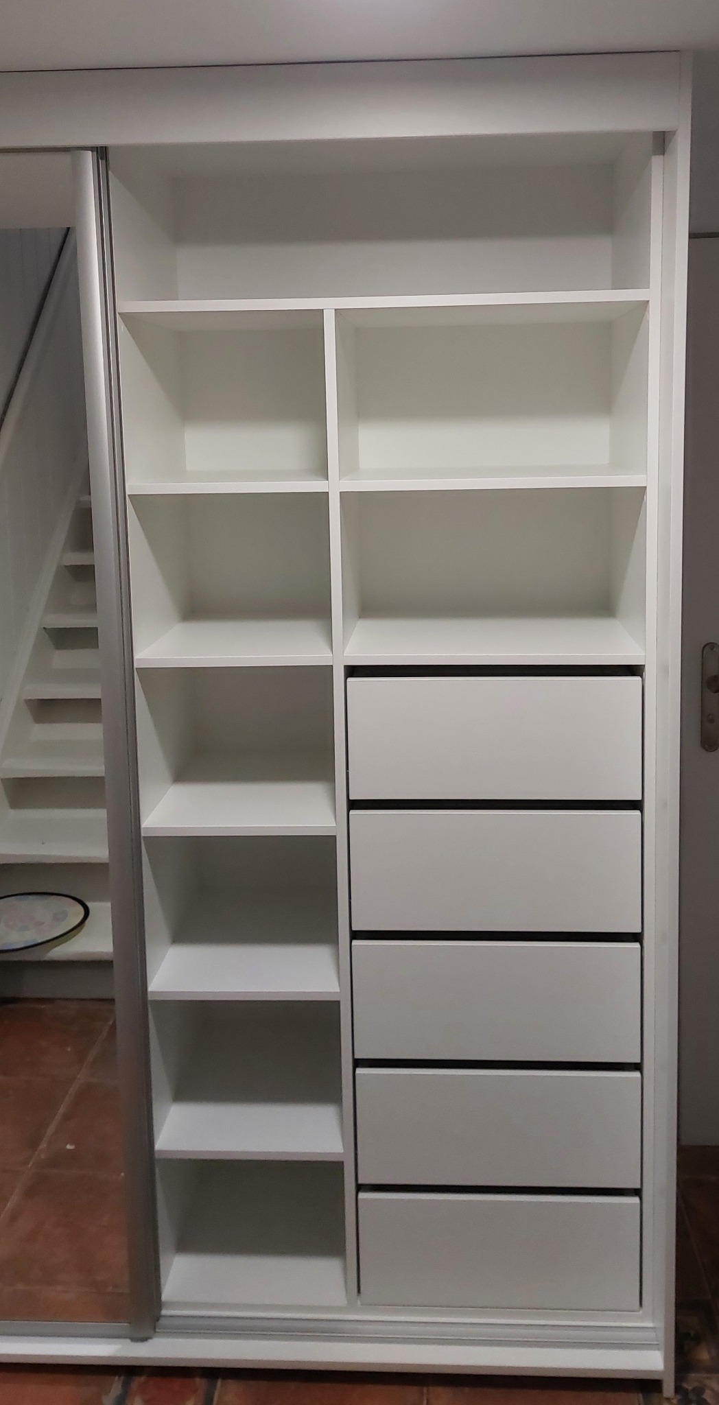 Biała szafa wnękowa z przesuwnymi drzwiami lustrzanymi, widoczne półki i szuflady z ciemnymi krawędziami, fragment schodów w odbiciu.