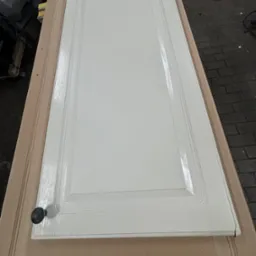 Białe, lakierowane drzwiczki szafki kuchennej z czarnym uchwytem, leżące na powierzchni roboczej z surowego drewna, prawdopodobnie po malowaniu lub renowacji.