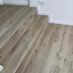 Ułożone panele podłogowe w odcieniach beżu z metalowymi listwami przejściowymi na schodach, białe listwy przypodłogowe i gniazdko elektryczne na białej, teksturowanej ścianie.