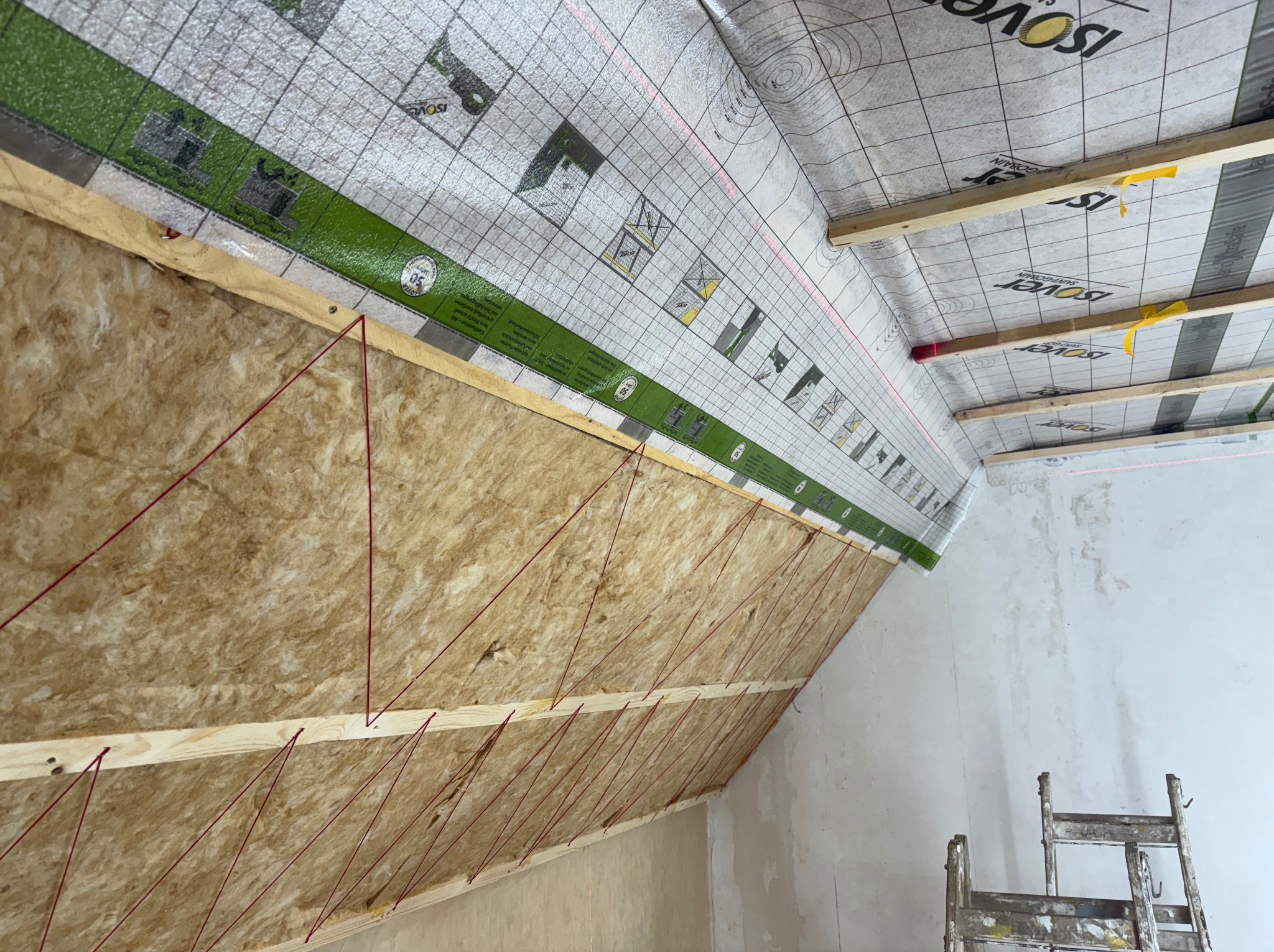 Izolacja termiczna poddasza z użyciem wełny mineralnej, widoczne drewniane legary, sznurki pomiarowe i folia paroizolacyjna z nadrukowaną siatką i logo Isover.