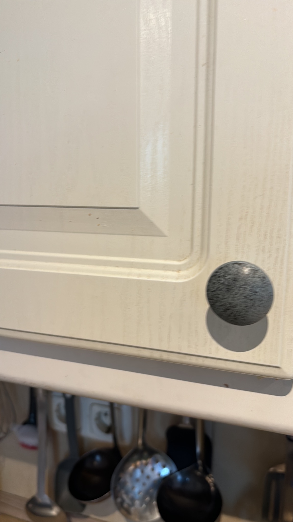 Biała szafka kuchenna z okrągłym, szarym uchwytem na tle zawieszonych przyborów kuchennych.