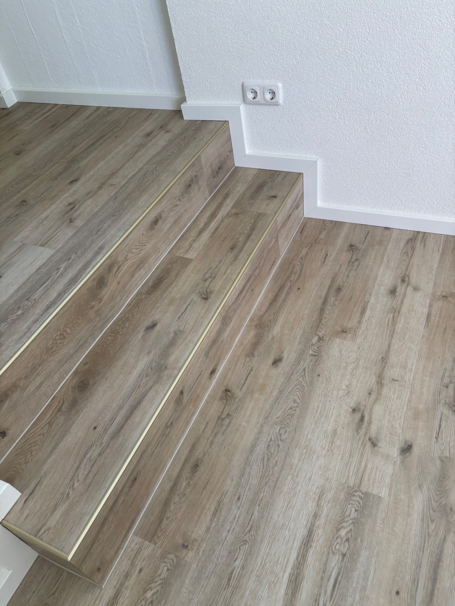 Ułożone panele podłogowe w odcieniach beżu z metalowymi listwami przejściowymi na schodach, białe listwy przypodłogowe i gniazdko elektryczne na białej, teksturowanej ścianie.