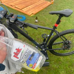 Dominik Mroczkowski - Czarny rower górski na trawie obok świeżo pomalowanych desek; w pierwszym planie butelka wosku samochodowego 'Plak Wosk na Mokro' trzymana w dłoni.