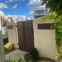 Nowoczesna brama z poziomych desek w jasnym ogrodzeniu, widok z zewnątrz na tle budynków mieszkalnych i błękitnego nieba z chmurami, z elementami zieleni.