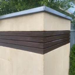 Fragment ogrodzenia z beżowego tynku z ciemnobrązowymi, drewnianymi panelami i metalowym daszkiem, otoczony zielenią.