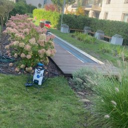 Dominik Mroczkowski - Myjka ciśnieniowa stojąca na trawie obok krzewu hortensji, drewnianej kładki i niskiego murku w otoczeniu nowoczesnej zabudowy osiedlowej.