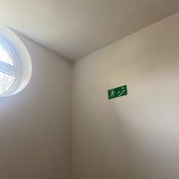 Dominik Mroczkowski - Wnętrze klatki schodowej z okrągłym oknem i zielonym znakiem ewakuacyjnym na ścianie, świeżo po malowaniu na jasny odcień.