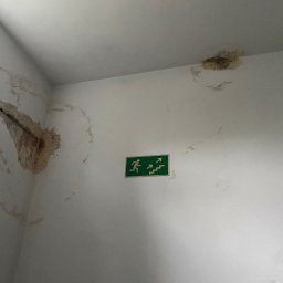 Dominik Mroczkowski - Zacieki na ścianie i suficie w rogu pomieszczenia, odsłonięty fragment zbrojenia, zielony znak ewakuacyjny.